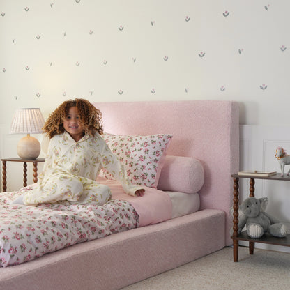 a girl sitting on a pink all cushioned bed bed with floral bedding.