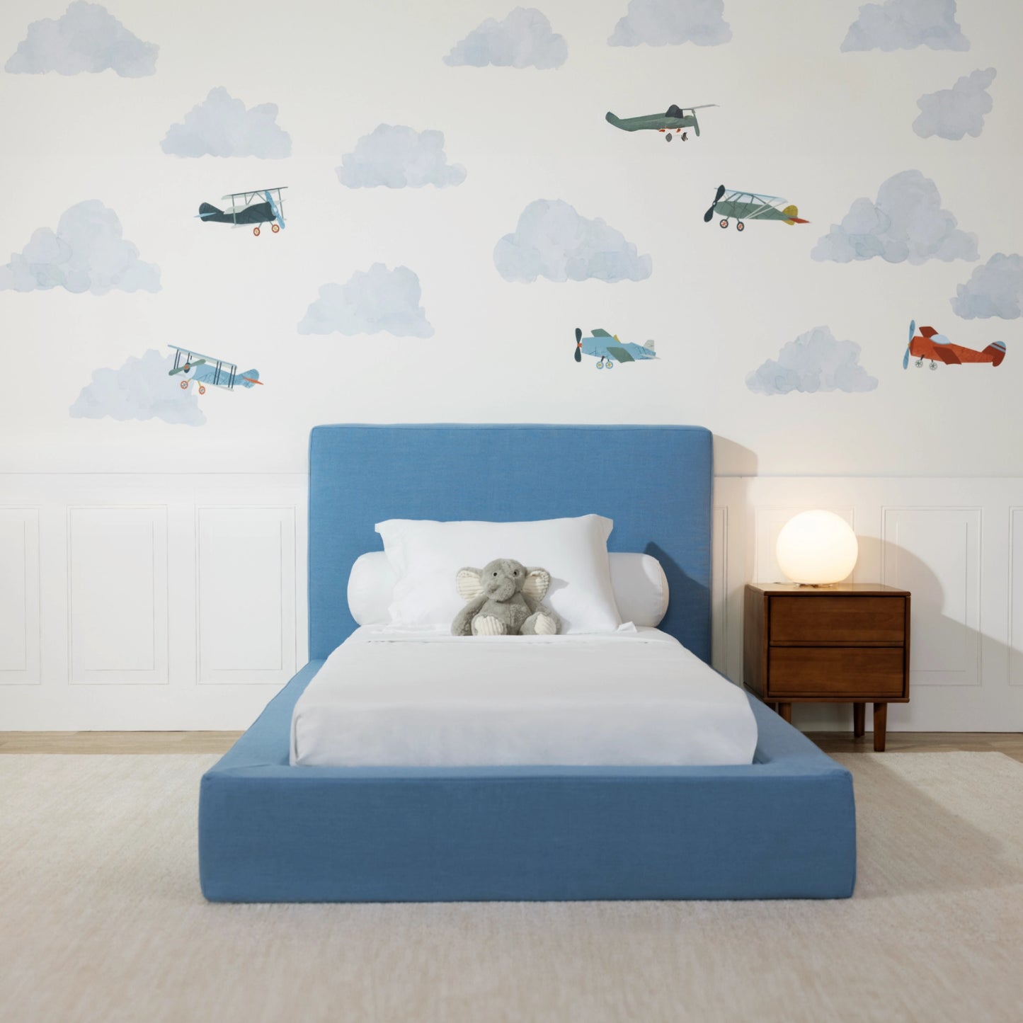 Front view of a blue all cushioned bed frame in a kids bed room with clouds and airplane decal. 