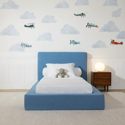 Front view of a blue all cushioned bed frame in a kids bed room with clouds and airplane decal. 