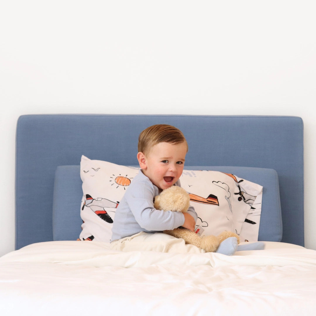 Child sitting on a all cushioned bed holding a teddy bear with a blue all cushioned headboard and airplane-patterned pillow.
