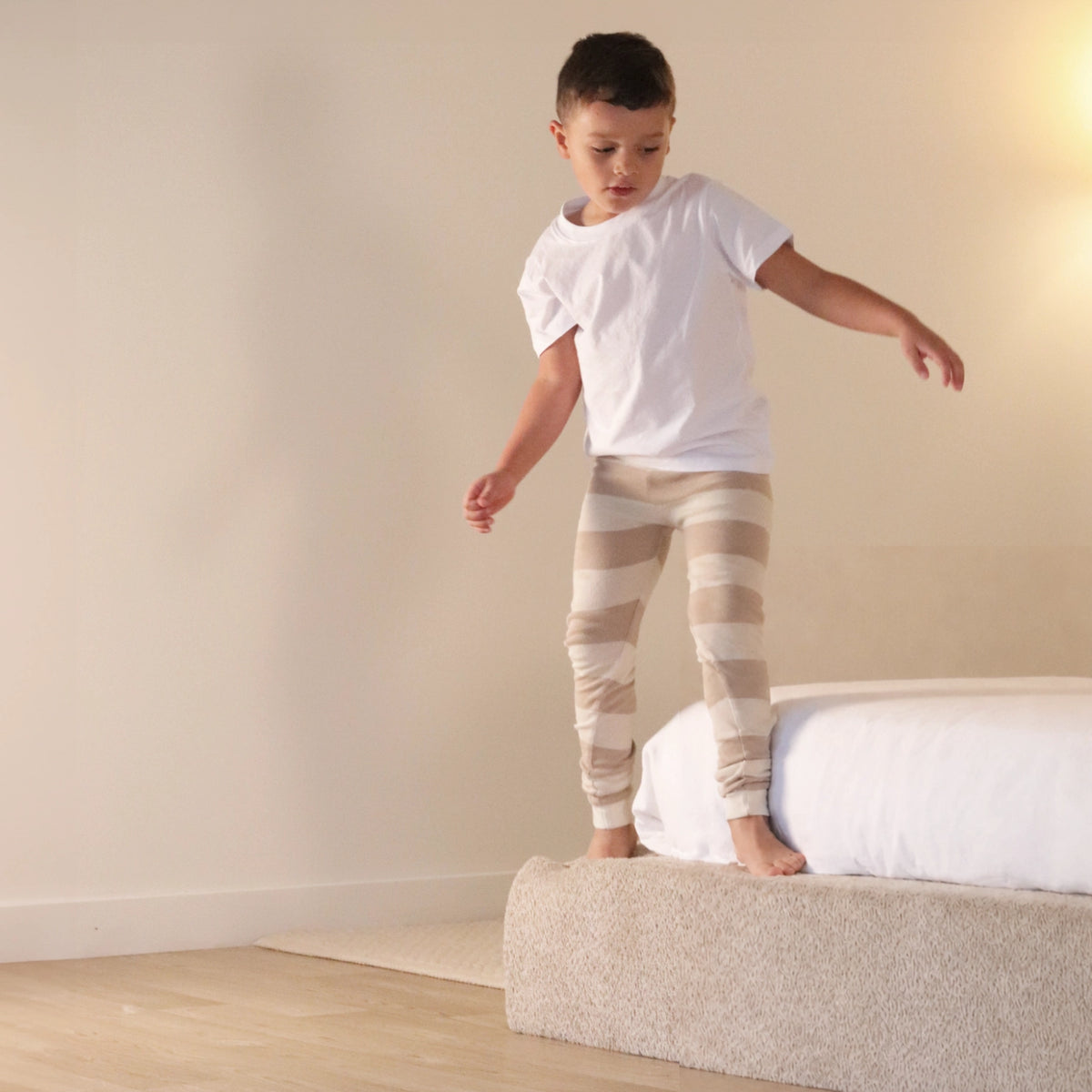 boy wearing a white t-shirt and striped pants standing on a all cushioned bed in a room with a plain wall.