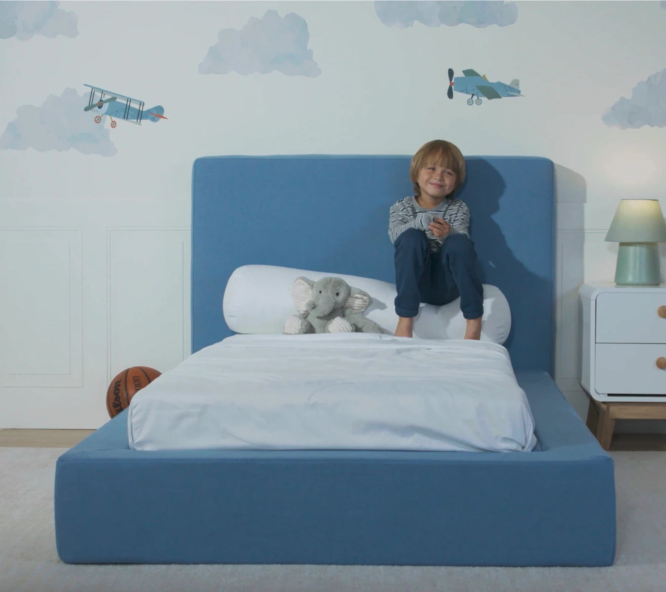 Child sitting on a blue bed with teddy bear, airplane decorations on wall | SoftFrame Kids