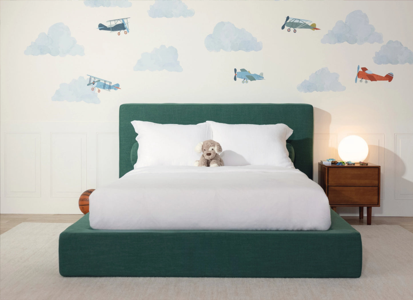 A modern children's bed with a dark green upholstered frame and headboard, dressed in white bedding, with a small plush dog on the mattress. The room features a white wall decorated with blue clouds and vintage airplanes, a wooden nightstand with a glowing globe lamp, and a light beige rug.