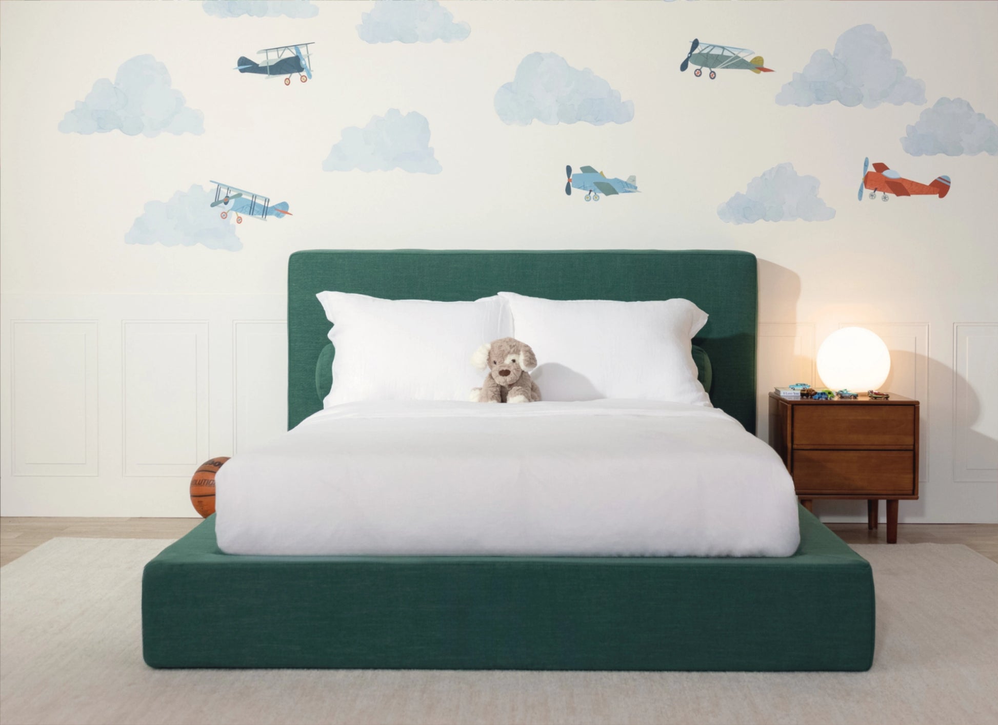 A modern children's bed with a dark green upholstered frame and headboard, dressed in white bedding, with a small plush dog on the mattress. The room features a white wall decorated with blue clouds and vintage airplanes, a wooden nightstand with a glowing globe lamp, and a light beige rug.