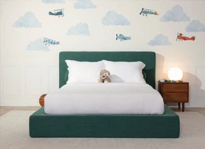 A modern children's bed with a dark green upholstered frame and headboard, dressed in white bedding, with a small plush dog on the mattress. The room features a white wall decorated with blue clouds and vintage airplanes, a wooden nightstand with a glowing globe lamp, and a light beige rug.